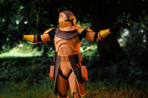 Ein Cosplayer in einem orangefarbenen, futuristischen Rüstungsoutfit mit zwei Pistolen, posiert vor einer grünen Kulisse. Die Szene fängt die kreative Atmosphäre des Elbenwald Festivals ein, das Musik und Nerdkultur vereint.