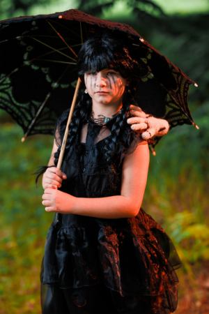 Ein Kind in einem schwarzen Kleid hält einen schwarzen Sonnenschirm und hat einen geheimnisvollen Ausdruck. Die Szenerie des Elbenwald Festivals vermittelt eine fantasievolle Atmosphäre, die Musik und Cosplay verbindet.