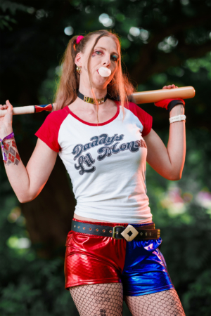 Frau in einem auffälligen Kostüm mit rotem und blauem glänzenden Shorts und einem T-Shirt, das "Daddy's Lil Monster" zeigt. Sie kaut Kaugummi und hält einen Baseballschläger über der Schulter. Hintergrund mit verschwommenem grünem Laub. Elbenwald Festival.