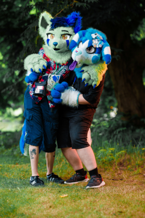 Zwei Personen in bunten Fursuits umarmen sich auf dem Elbenwald Festival, umgeben von grüner Natur. Die kreative Kostümierung zeigt eine Mischung aus tierischen Elementen und farbenfrohen Designs, die die lebendige Atmosphäre des Festivals widerspiegeln.