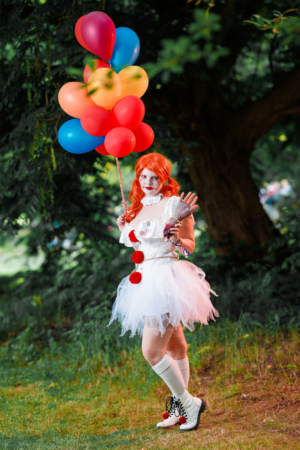 Eine Person in einem weißen Tutu-Kleid mit roten Pompons und roten Haaren hält bunte Luftballons. Die Szene ist lebhaft und zeigt die kreative Kostümkultur des Elbenwald Festivals, das Musik und Fantasie verbindet.