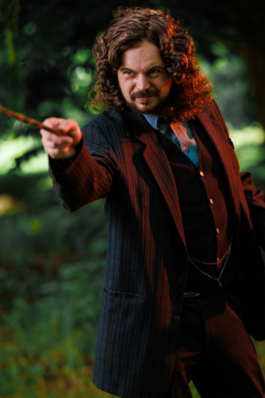 Ein männlicher Cosplayer mit lockigem Haar und einem gestreiften Anzug hält einen Zauberstab in der Hand. Er posiert im Freien, umgeben von einer grünen Kulisse, und vermittelt eine magische Atmosphäre, die zur nerdigen Stimmung des Elbenwald Festivals passt.