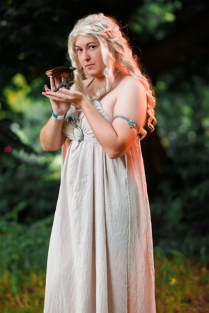 Kostümierte Person mit blonden Haaren und einem hellen Kleid hält eine kleine Drachenfigur in der Hand. Die Szene spielt im Freien, umgeben von grüner Natur, und spiegelt das kreative und fantasievolle Ambiente des Elbenwald Festivals wider.