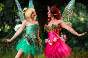 Zwei Frauen in bunten Feen-Kostümen mit funkelnden Flügeln posieren fröhlich auf dem Elbenwald Festival, umgeben von üppiger Natur. Die eine trägt ein grünes, die andere ein pinkes Kleid, beide geschmückt mit Blumen und Pflanzen.