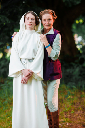 Zwei Frauen in Kostümen, inspiriert von einer bekannten Science-Fiction-Serie, posieren lächelnd im Freien. Die erste trägt ein weißes Gewand mit Kapuze, die zweite ein lila Oberteil und braune Stiefel. Hintergrund mit grüner Natur, typisch für Festivals.