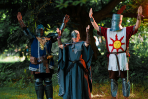 Drei kostümierte Teilnehmer des Elbenwald Festivals posieren im Freien. Sie tragen mittelalterliche Rüstungen und Gewänder, während sie ihre Arme heben. Die Szene vermittelt eine festliche Atmosphäre, die die Verbindung von Musik und Fantasie betont.