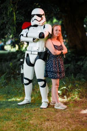 Ein Stormtrooper aus Star Wars steht neben einer Frau in einem blumigen Kleid auf dem Elbenwald Festival. Die Szene zeigt die Verbindung von Musik und Nerd-Kultur in einer lebhaften Festivalatmosphäre im Freien.