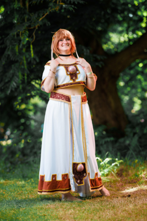 Eine Cosplayerin in einem eleganten, weißen Kostüm mit goldenen und roten Akzenten, das an ein Fantasy-Charakterdesign erinnert. Sie posiert fröhlich im Freien, umgeben von grüner Natur, während das Licht sanft auf sie fällt. Elbenwald Festival 2023.