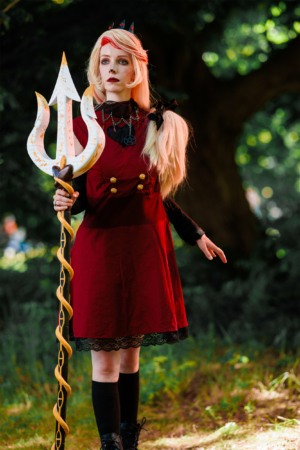 Eine kostümierte Person mit blonden Haaren und einem roten Kleid hält einen stilisierten Dreizack. Der Hintergrund ist grün und natürlich, was die lebendige Atmosphäre des Elbenwald Festivals unterstreicht.