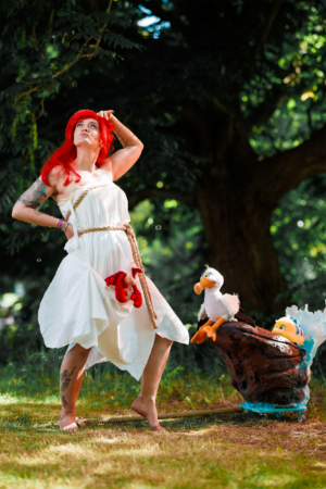 Cosplay einer Figur mit rotem Haar in einem weißen Kleid, umgeben von plüschigen Meerestieren, während sie in einer natürlichen Umgebung steht. Die Szene verkörpert die kreative Atmosphäre des Elbenwald Festivals, das Musik und Nerdkultur vereint.