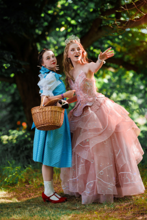 Zwei Frauen in fantasievollen Kostümen stehen im Freien: eine in einem blauen Kleid mit einem Korb und die andere in einem glitzernden rosa Kleid mit einer Krone. Sie scheinen sich auf dem Elbenwald Festival zu unterhalten und die Umgebung zu erkunden.
