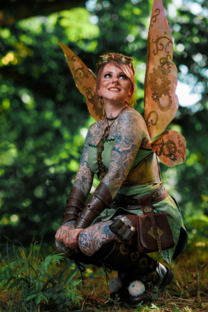 Eine verkleidete Frau mit bunten Schmetterlingsflügeln und Tattoos, die in der Natur sitzt. Ihr Outfit kombiniert Steampunk-Elemente mit einem fantasievollen Look, ideal für das Elbenwald Festival, das Musik und Nerdkultur vereint.