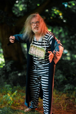 Ein kostümierter Teilnehmer des Elbenwald Festivals in einem gestreiften Gefängniskostüm mit einem Schild, das "Azkaban Prison" zeigt, posiert im Freien. Die Szenerie ist von natürlichem Grün umgeben, was die fantasievolle Atmosphäre des Festivals unterstreicht.