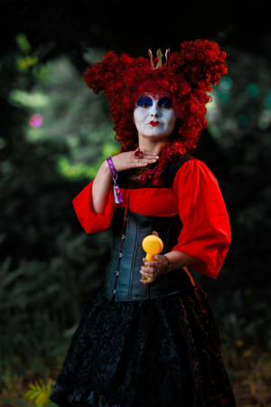 Eine künstlerisch verkleidete Person mit rotem Haar und bunter Schminke posiert beim Elbenwald Festival. Sie trägt ein schwarzes Korsett und einen langen Rock, während sie einen gelben Gegenstand in der Hand hält, umgeben von einer lebhaften Festivalatmosphäre.