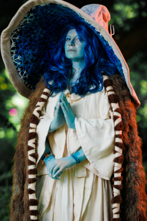 Eine kostümierte Person mit blauer Haut und langen blauen Haaren steht mit gefalteten Händen da. Sie trägt einen langen, hellen Umhang mit einer fellartigen Verzierung und einem großen, bunten Hut. Der Hintergrund zeigt eine grüne, natürliche Umgebung.