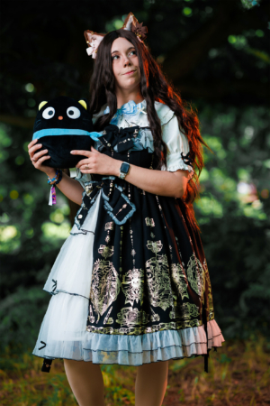 Ein Festivalbesucher trägt ein elegantes, schwarzes Lolita-Kleid mit goldenen Verzierungen und hält ein schwarzes Plüschtier. Die Szenerie ist von grünen Bäumen umgeben, was eine lebendige und nostalgische Atmosphäre schafft, die zur nerdigen Kultur des Elbenwald Festivals passt.