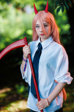 Cosplay eines Mädchens mit rosa Haaren und roten Teufelshörnern, das eine rote Sense hält. Sie trägt ein weißes Hemd mit schwarzer Krawatte und steht vor einer grünen Hintergrundlandschaft beim Elbenwald Festival, einem Event für Nerds und Fans.