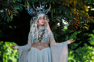 Kostümierte Person mit einem beeindruckenden Geweih-Haarschmuck und einem fließenden, grauen Gewand, steht vor einem grünen Hintergrund beim Elbenwald Festival, das Musik und Fantasy verbindet. Die Atmosphäre ist magisch und kreativ.