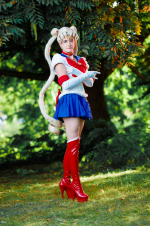 Cosplayer auf dem Elbenwald Festival, verkleidet als Sailor Moon mit langen blonden Haaren, rotem Oberteil und blauer Minirock. Sie trägt hohe, rote Stiefel und posiert vor einer grünen Kulisse mit Bäumen. Ideal für Fans von Anime und Cosplay.