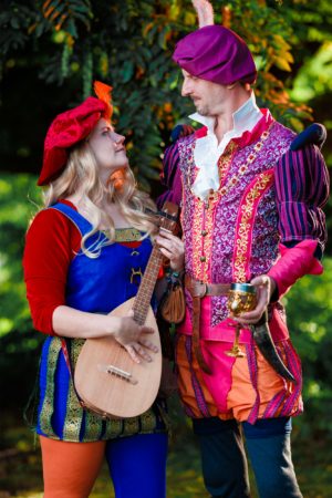 Zwei kostümierte Festivalbesucher auf dem Elbenwald Festival, die sich anlächeln. Die Frau trägt ein buntes Outfit und hält eine Laute, während der Mann ein prunkvolles Gewand und einen goldenen Becher in der Hand hält. Grüner Hintergrund sorgt für eine festliche Atmosphäre.