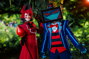 Kreative Cosplay-Darstellung auf dem Elbenwald Festival: Zwei Personen in farbenfrohen Kostümen, einer mit rotem Outfit und Stab, der andere mit einem Bildschirmkopf. Im Hintergrund ist eine grüne Umgebung zu sehen, die die festliche Atmosphäre unterstreicht.