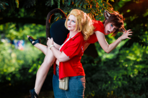 Zwei Frauen in Kostümen auf dem Elbenwald Festival: Eine trägt ein rotes Oberteil und hat lange blonde Haare, während die andere mit einem tierischen Look und Ohren posiert. Die Szene ist lebhaft und zeigt die kreative Atmosphäre des Festivals im Freien.