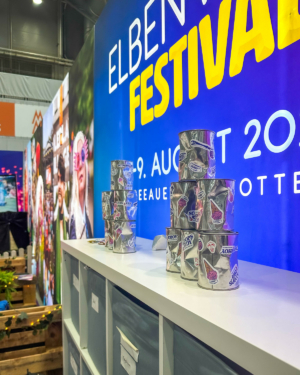 Stapel aus bunten Eiskübel-Dosen auf einem Tisch vor dem farbenfrohen Hintergrund des Elbenwald Festivals. Die Veranstaltung verbindet Musik und Nerd-Kultur und findet im August 2023 in Göttingen statt.