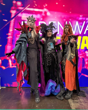 Drei Cosplayer in beeindruckenden, fantasievollen Kostümen stehen vor einem bunten Hintergrund beim Elbenwald Festival. Die Outfits sind von dunkler Magie inspiriert und zeigen kreative Details, die die Besucher begeistern.