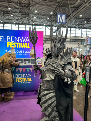 Ein beeindruckender Cosplayer in einer detaillierten Rüstung, inspiriert von Fantasy-Charakteren, posiert am Eingang des Elbenwald Festivals. Im Hintergrund ist das Festival-Logo und bunte Stände sichtbar, die die nerdige Atmosphäre unterstreichen.