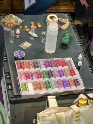 Bunte Glitzerstäube in durchsichtigen Behältern auf einem Tisch, umgeben von Bastelmaterialien wie Flaschen, Kleber, Korken und Garn. Ideal für kreative Projekte auf dem Elbenwald Festival, das Musik und Nerd-Kultur vereint.
