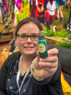 Eine Frau mit Brille hält stolz ein kleines Fläschchen mit grünem Inhalt und einem Anhänger in der Hand. Im Hintergrund sind bunte Festivalbesucher zu sehen, die zur lebhaften Atmosphäre des Elbenwald Festivals passen.