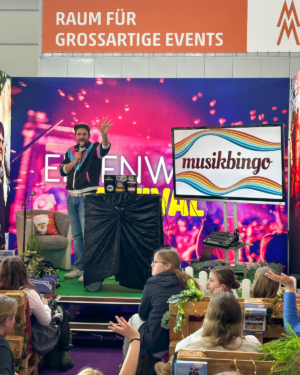 Ein Moderator präsentiert Musikbingo auf der Bühne des Elbenwald Festivals. Im Hintergrund sind bunte Lichter und eine große Leinwand zu sehen. Das Publikum sitzt auf Holzpaletten und ist gespannt auf das Event.