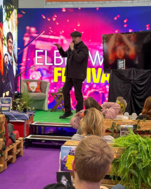 Ein Sprecher steht auf der Bühne des Elbenwald Festivals und spricht in ein Mikrofon, während das Publikum aufmerksam zuhört. Im Hintergrund leuchtet ein farbenfrohes Festival-Design. Die Atmosphäre ist lebhaft und einladend für Fans von Musik und Nerd-Kultur.