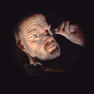 Ein älterer Mann mit Brille und Bart schaut neugierig in die Kamera. Sein Gesichtsausdruck ist intensiv und nachdenklich. Der dunkle Hintergrund verstärkt die mysteriöse Atmosphäre, die zur nerdigen Stimmung des Elbenwald Festivals passt.