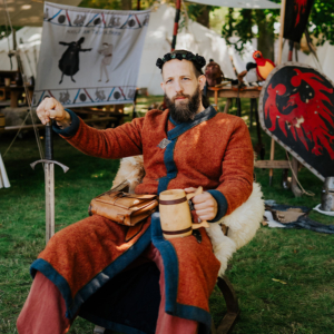 Ein Mann in historischer Kleidung sitzt auf einem Stuhl und hält ein Trinkgefäß sowie ein Schwert. Im Hintergrund sind Zelte und dekorative Elemente des Elbenwald Festivals zu sehen, das Musik und Nerd-Kultur vereint.