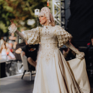 Eine Person in einem eleganten, weißen Kostüm mit Rüschen und Perlen steht auf der Bühne des Elbenwald Festivals, umgeben von Zuschauern. Die Atmosphäre ist lebhaft und kreativ, ideal für Fans von Musik und Popkultur.