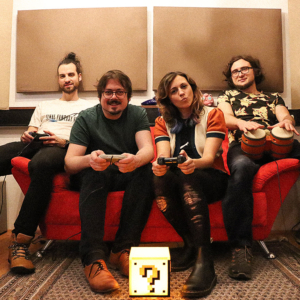 Endgegner Vier Personen sitzen auf einem roten Sofa, spielen Videospiele mit Controllern und genießen die Zeit zusammen. Vor ihnen steht eine leuchtende Fragezeichen-Box, die an klassische Spiele erinnert. Ein gemütliches, nerdiges Ambiente, perfekt für das Elbenwald Festival.