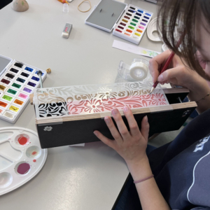 Eine Person bemalt mit Pinsel und Farben eine dekorative Holzbox, umgeben von Malutensilien, während sie am Elbenwald Festival kreativ arbeitet. Die Szene zeigt die Verbindung von Kunst und handwerklicher Gestaltung in einem inspirierenden Umfeld.
