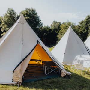 Zelt im Freien beim Elbenwald Festival, umgeben von Bäumen. Das Zelt hat eine offene Eingangstür, die einen Blick auf zwei Schlafplätze im Inneren bietet. Rasenfläche und weitere Zelte im Hintergrund schaffen eine einladende Festivalatmosphäre.