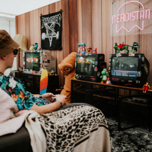 Ein Besucher sitzt auf einem Sofa im Nerdistan-Bereich des Elbenwald Festivals, umgeben von Retro-Fernsehern, Spielzeugfiguren und einem bunten Skateboard. Die entspannte Atmosphäre lädt zum Spielen und Verweilen ein, während ein neonfarbener Schriftzug den Raum beleuchtet.