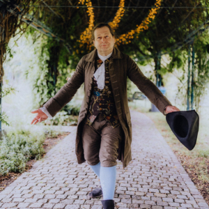 Ein kostümiertes Festivalbesucher in historischer Kleidung mit Hut, der auf einem gepflasterten Weg unter einer mit Lichtern geschmückten Laube beim Elbenwald Festival spaziert. Die Atmosphäre ist festlich und einladend, ideal für Fans von Musik und Fantasy.