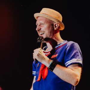 Olaf Unzel, Foto: Rainer Keuenhof Ein lächelnder Mann in einem maritimen Kostüm mit Strohhut hält ein Mikrofon auf der Bühne des Elbenwald Festivals, umgeben von bunten Lichtern. Die Atmosphäre ist festlich und energiegeladen, ideal für Musik und Nerd-Kultur.