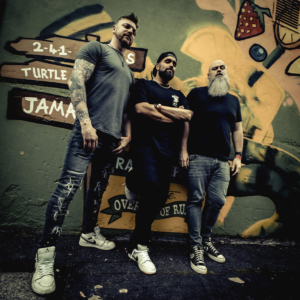 Drei Männer posieren vor einer bunten Wand mit Graffiti beim Elbenwald Festival. Die Szene strahlt eine lässige Atmosphäre aus, die die Verbindung von Musik und Nerdkultur widerspiegelt. Perfekt für Fans von alternativer Unterhaltung und Festivals.