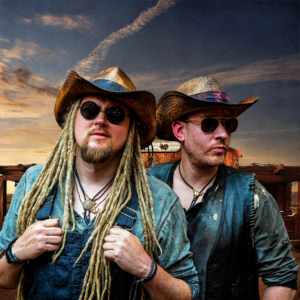 Zwei Männer im Western-Stil mit Cowboyhüten und Sonnenbrillen posieren vor einer malerischen Kulisse. Ihre Outfits und Accessoires spiegeln das kreative Flair des Elbenwald Festivals wider, das Musik und Nerd-Kultur verbindet.