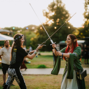 Zwei Frauen im Cosplay kämpfen mit Lichtschwertern auf dem Elbenwald Festival. Die Szene zeigt eine lebendige Atmosphäre mit bunten Kostümen und fröhlichen Teilnehmern im Hintergrund, die die Mischung aus Musik und Nerdkultur genießen.