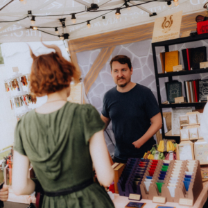 Ein Stand auf dem Elbenwald Festival, wo ein Verkäufer mit einem interessierten Kunden spricht. Im Hintergrund sind farbige Notizbücher und Merchandise-Artikel sichtbar, die die kreative Atmosphäre des Festivals widerspiegeln.