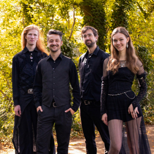 Vier Musiker posieren in stilvollen, dunklen Outfits vor einer grünen Kulisse beim Elbenwald Festival, einem einzigartigen Event, das Musik und Nerdkultur vereint. Die Atmosphäre ist lebhaft und kreativ, ideal für Fans von Fantasy und Musik.