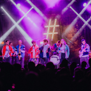 #zweiraumsilke, Foto: Rainer Keuenhof Bühne beim Elbenwald Festival mit einer Band, die in bunten Outfits auftritt. Die Musiker spielen verschiedene Instrumente vor einem begeisterten Publikum, umgeben von farbenfrohen Lichtern und einer lebhaften Atmosphäre.